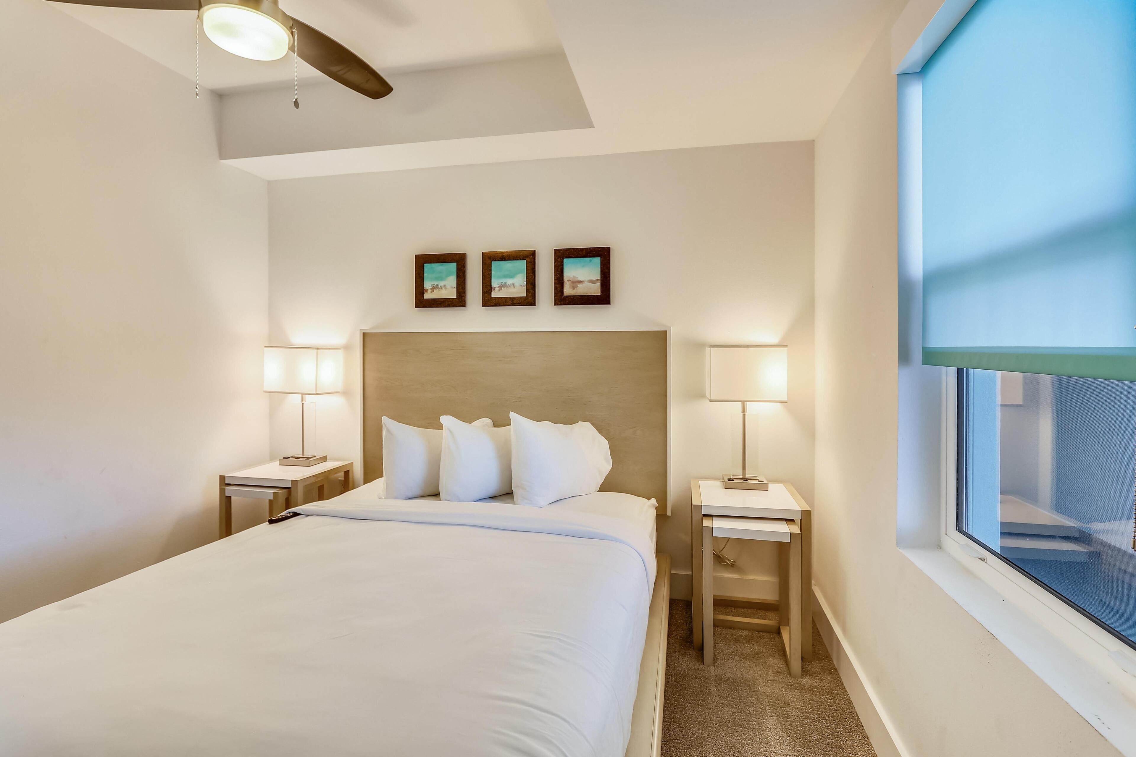 Una habitación de hotel con una cama, dos mesitas de noche con lámparas, un ventilador de techo y una ventana.