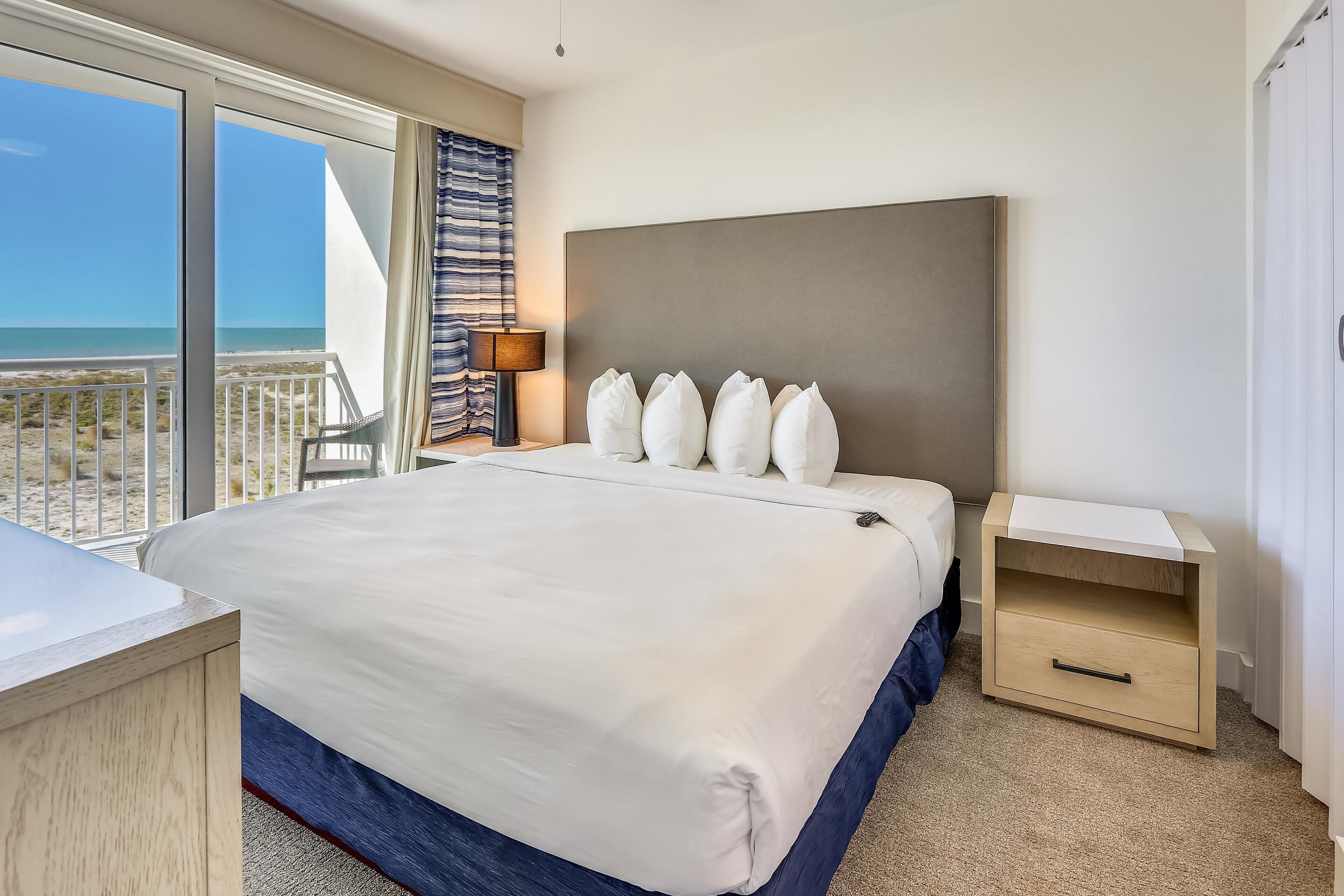 Ein Hotelzimmer mit einem großen Bett, einem Nachttisch, einem Schreibtisch und Blick auf den Strand.