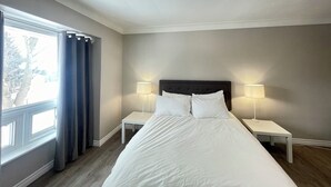 A hotel room with a large bed, two bedside tables with lamps, a window with curtains, and a ceiling light fixture. - Mountain View Suite - Sleeps 6
 (St. Catharines)