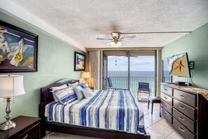 Relax in this serene hotel room with a large bed, dark dresser, and comfortable chair by the window, offering a view of the ocean from the balcony.