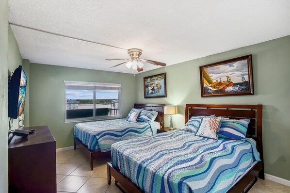 Two single beds with blue and white striped bedding, a ceiling fan, and a large window offering a view of the outside.