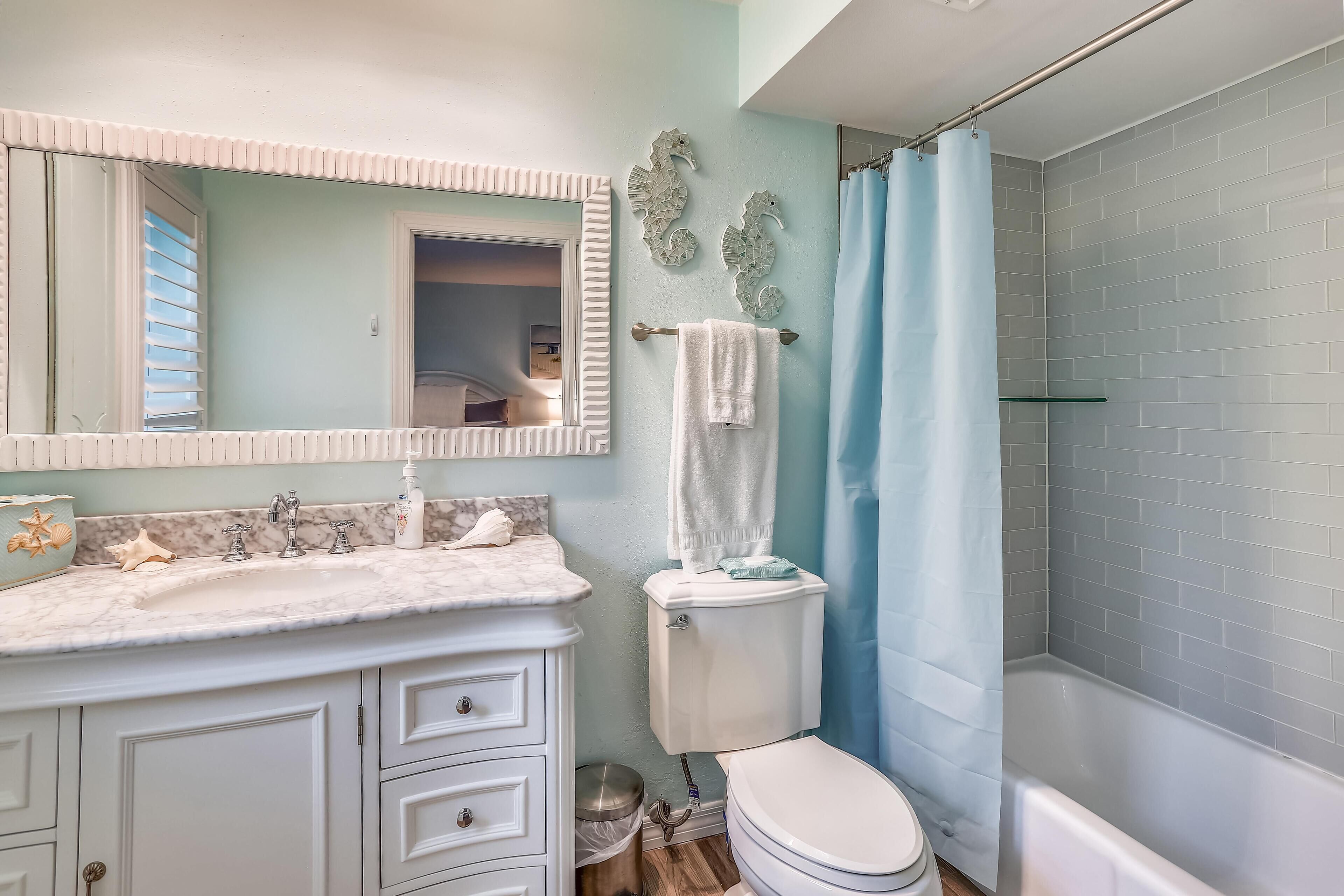 Een badkamer met een witte wastafel, een toilet, een douche met een blauw gordijn en een spiegel met decoratieve zeepaard muurdecoratie.
