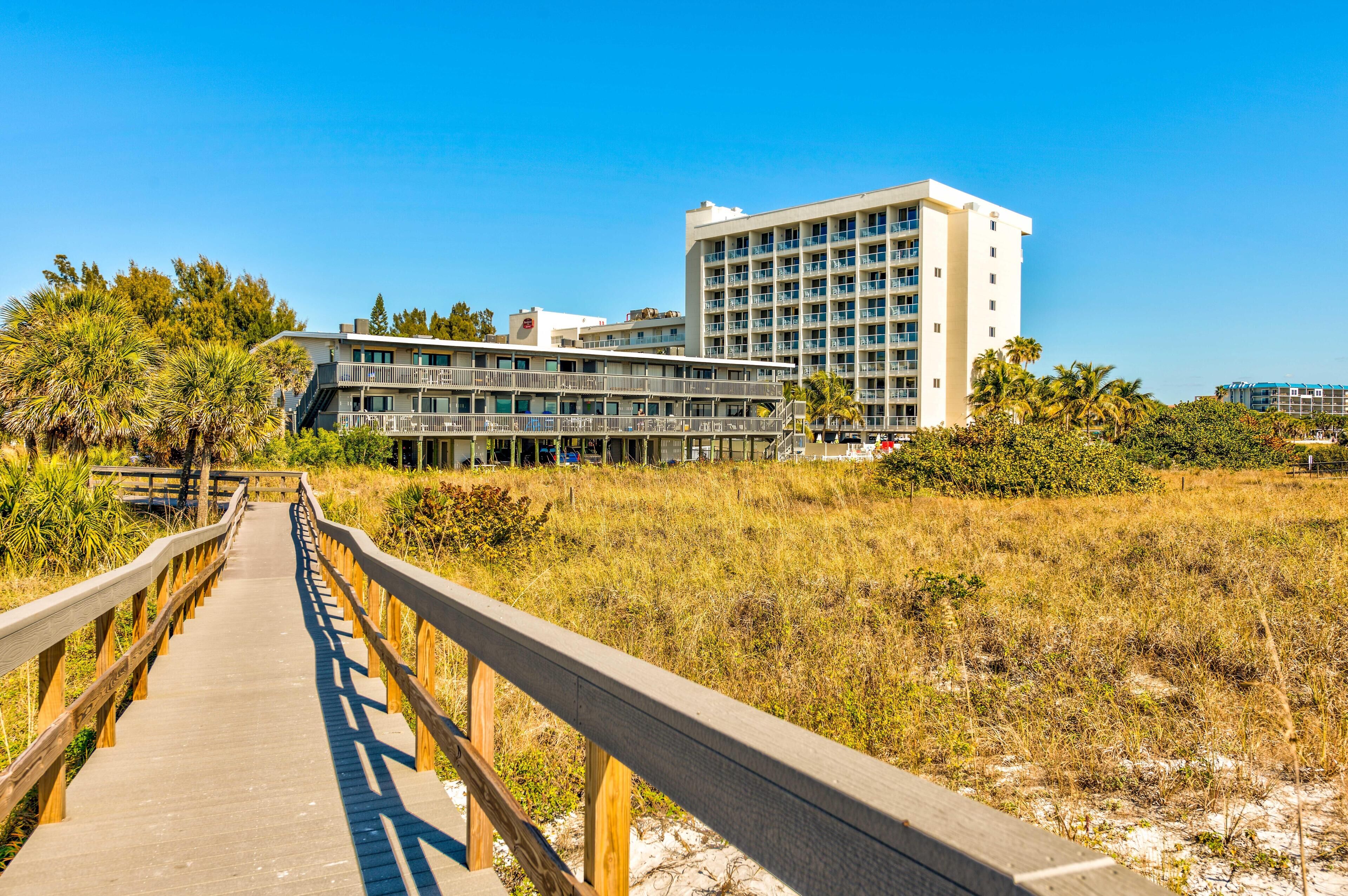 A wooden boardwalk leading to a hotel with multiple floors and balconies, surrounded by natural vegetation.