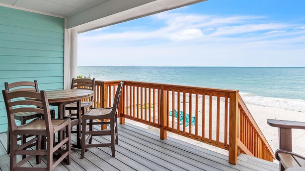 A wooden deck with a table and chairs, overlooking a beach with clear blue water and a wooden railing. - Serenity By The Sea (Treasure Island)