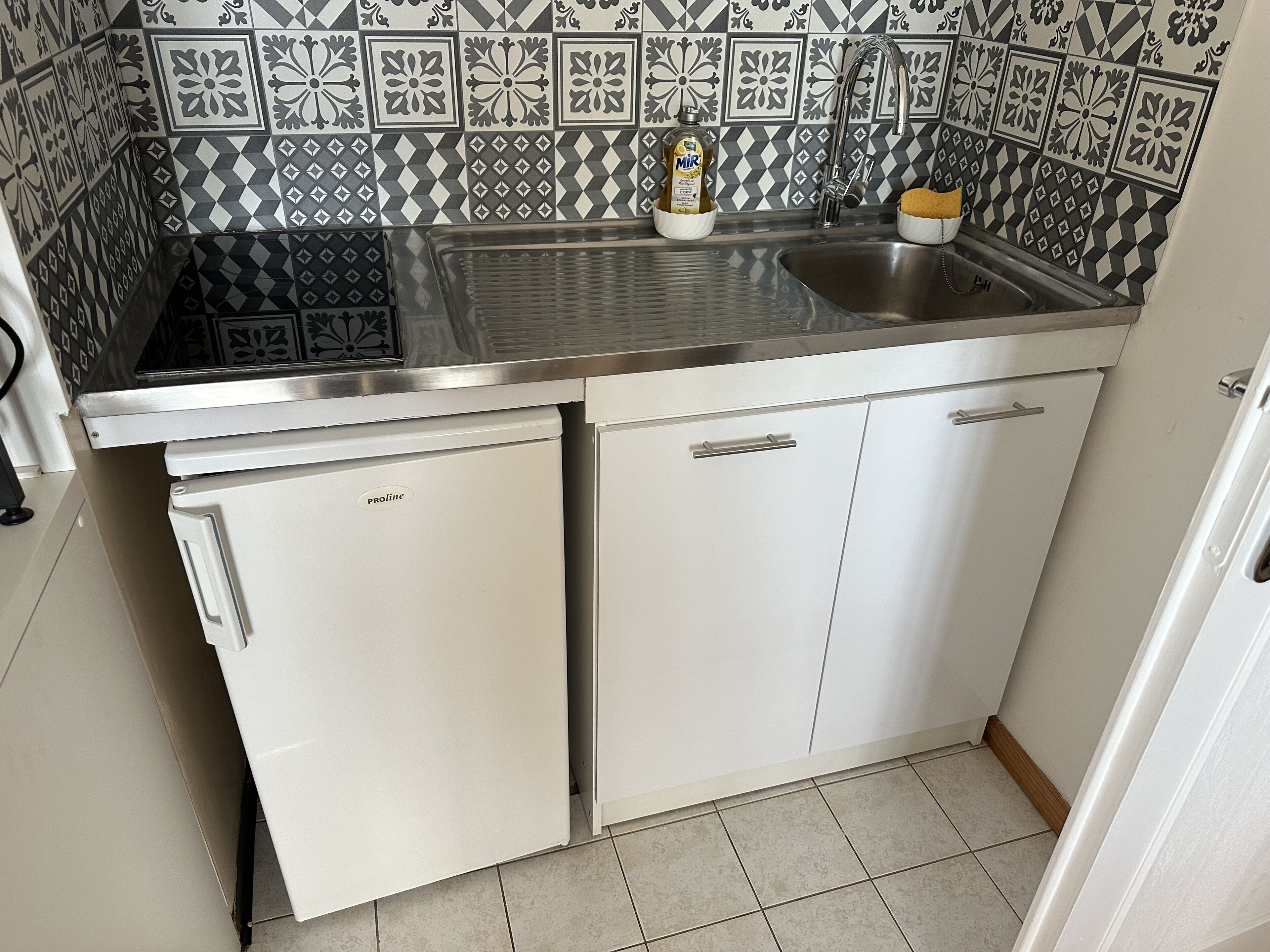 A compact kitchen with a stainless steel sink, a white refrigerator, and a patterned backsplash.