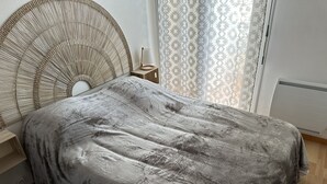 A bedroom with a grey textured bedspread, a woven headboard, a bedside lamp, and patterned curtains. - Appartement Les Lupins vue Port (Courseulles-sur-Mer)