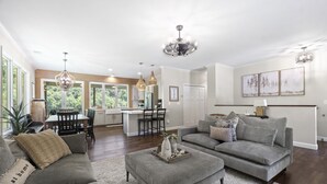 Ein großzügiges Wohnzimmer mit grauem Sofa, Hocker und einem Essbereich mit Tisch und Stühlen.