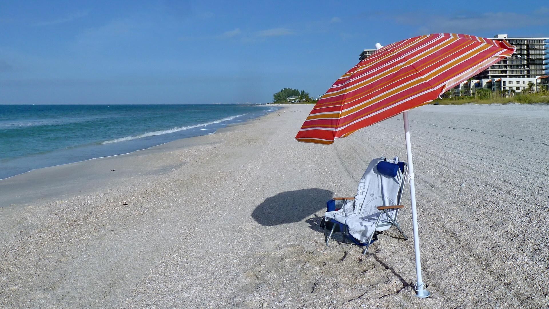 Une plage avec un parasol rayé, une chaise de plage et un bâtiment en arrière-plan.