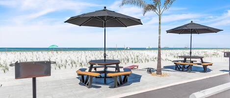 A beachside picnic area with tables and umbrellas, a palm tree, and a view of the ocean.