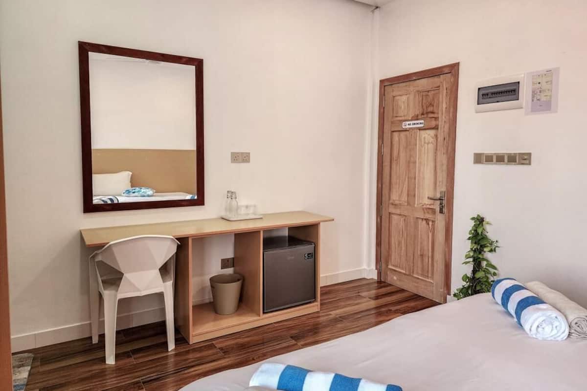 Ett hotellrum med en säng, ett skrivbord med en stol, ett litet kylskåp, en trädörr och en växt.