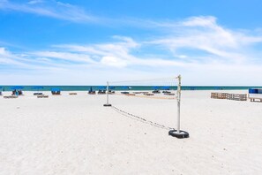 Uma rede de voleibol de praia numa praia arenosa, com guarda-sóis azuis e bancos de madeira.