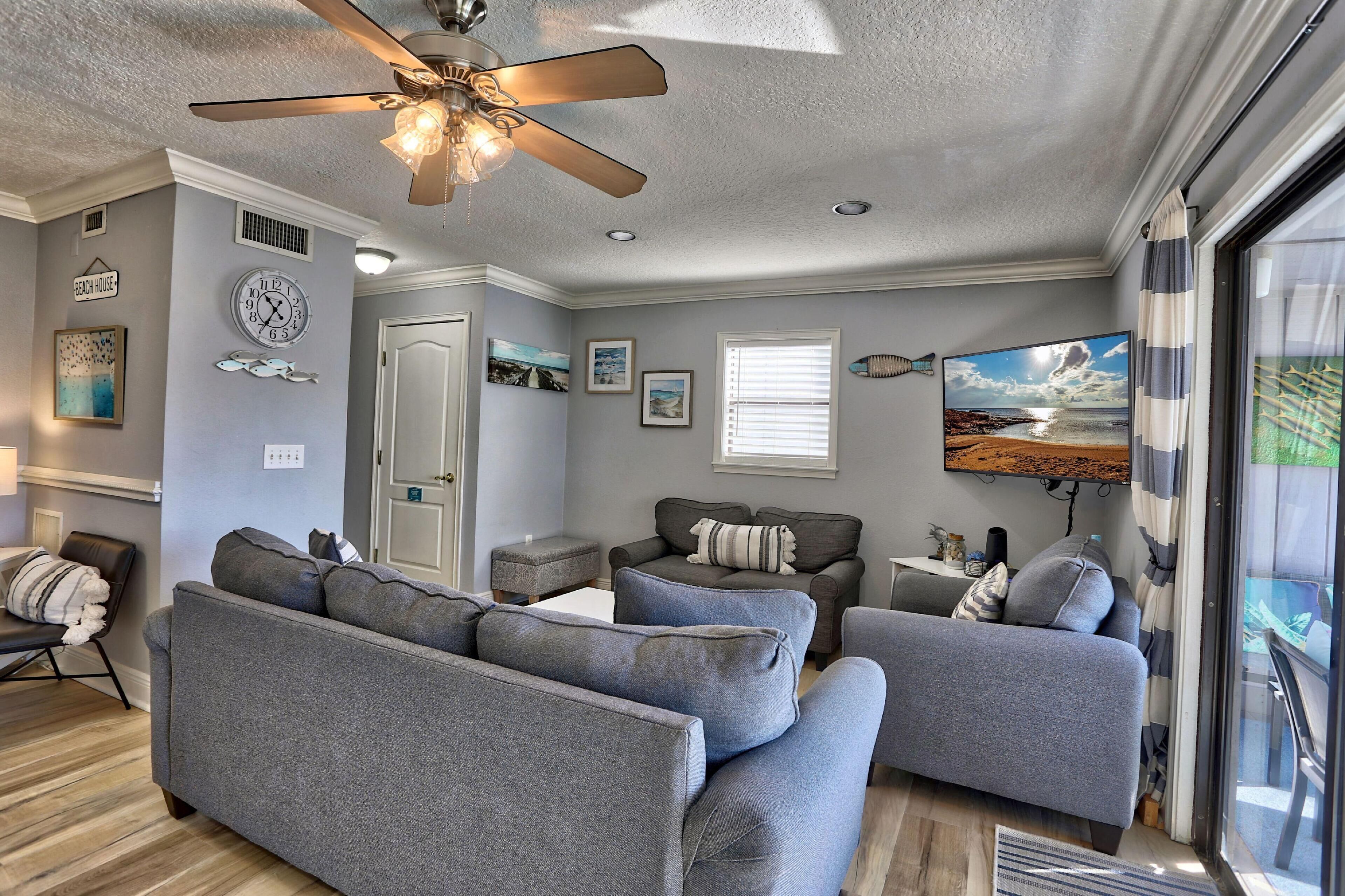 A modern living room with grey sofas, a ceiling fan, and a large window with blinds.