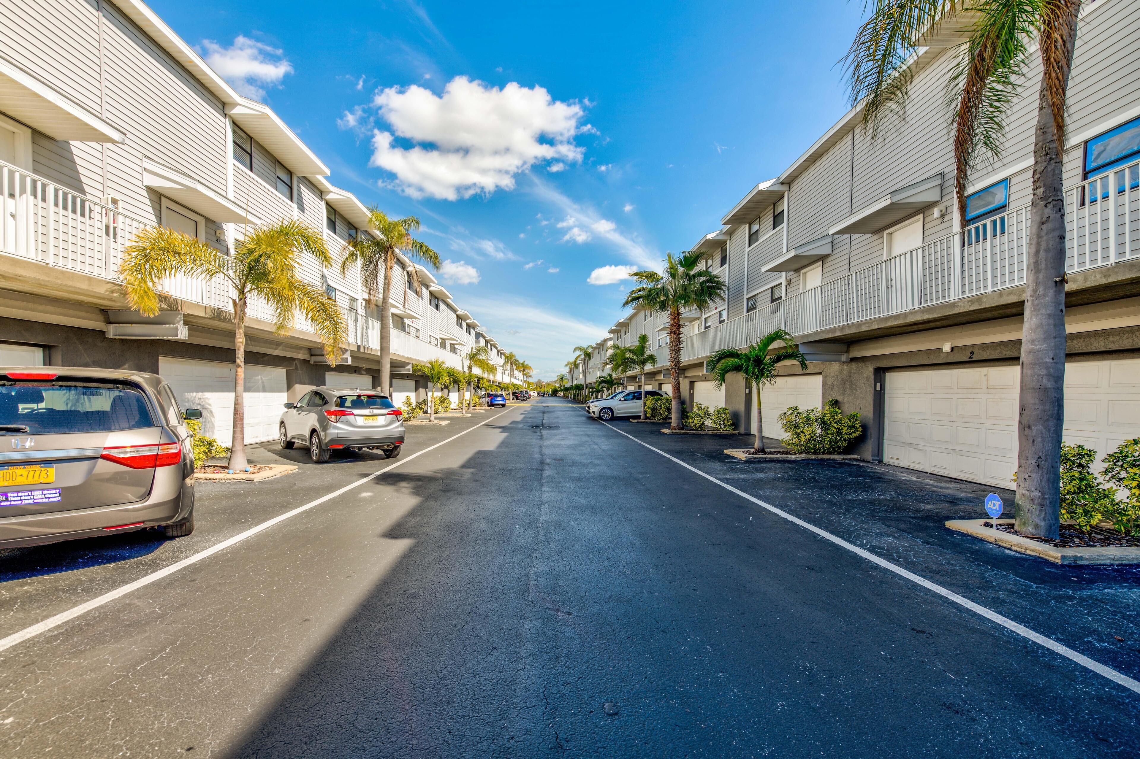 A residential street with multi-story houses, parked cars, and palm trees.