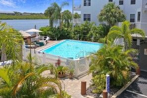 A swimming pool surrounded by palm trees and a white fence, with a view of a body of water and a building in the background. - Sea Shell 24 (Indian Shores)