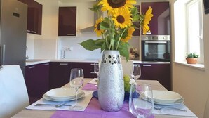 A dining area with a table set for two, a vase with sunflowers, and a kitchen in the background.
