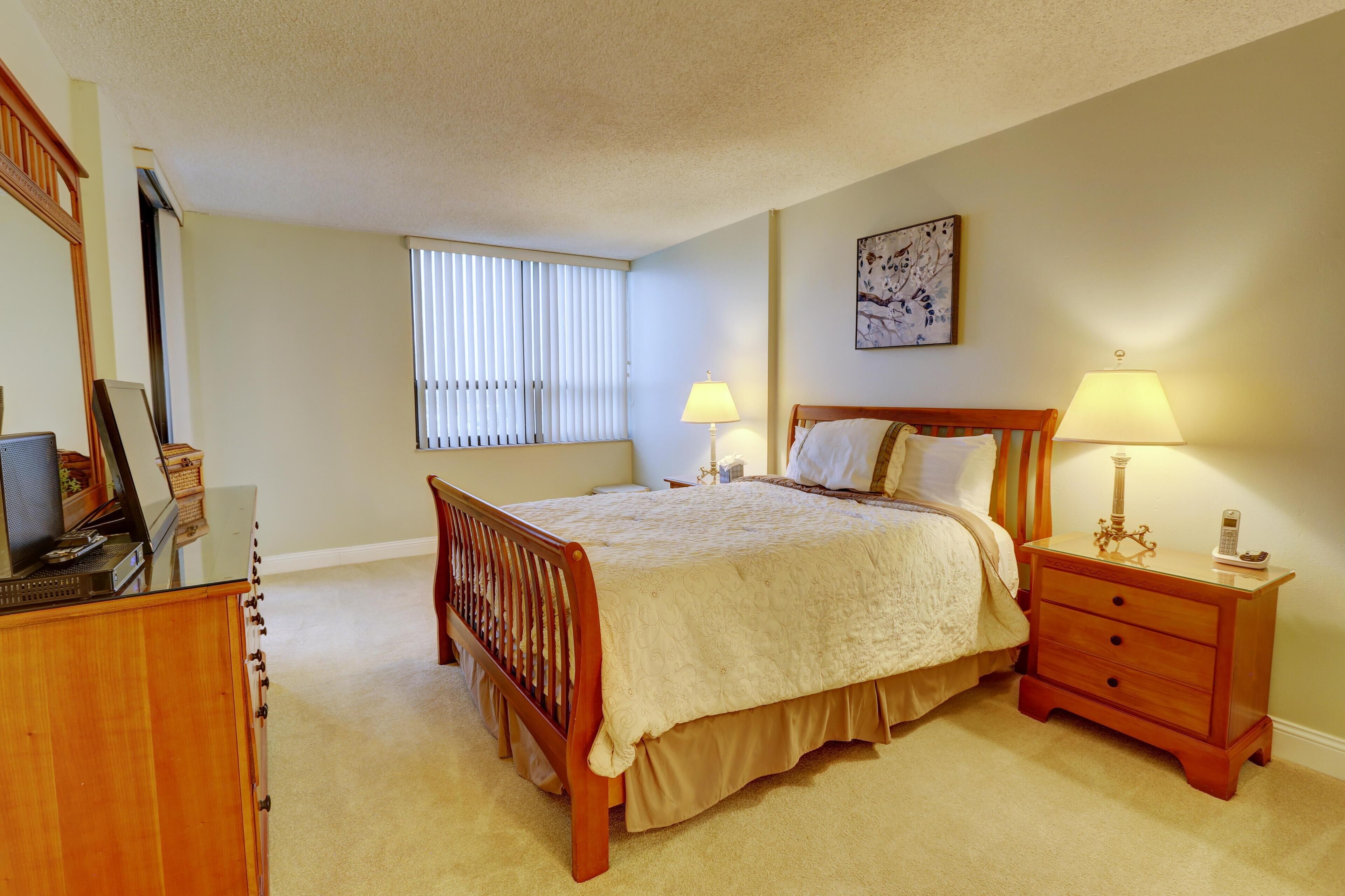 A bedroom with a bed, bedside tables, a dresser, a window with blinds, and a painting on the wall.