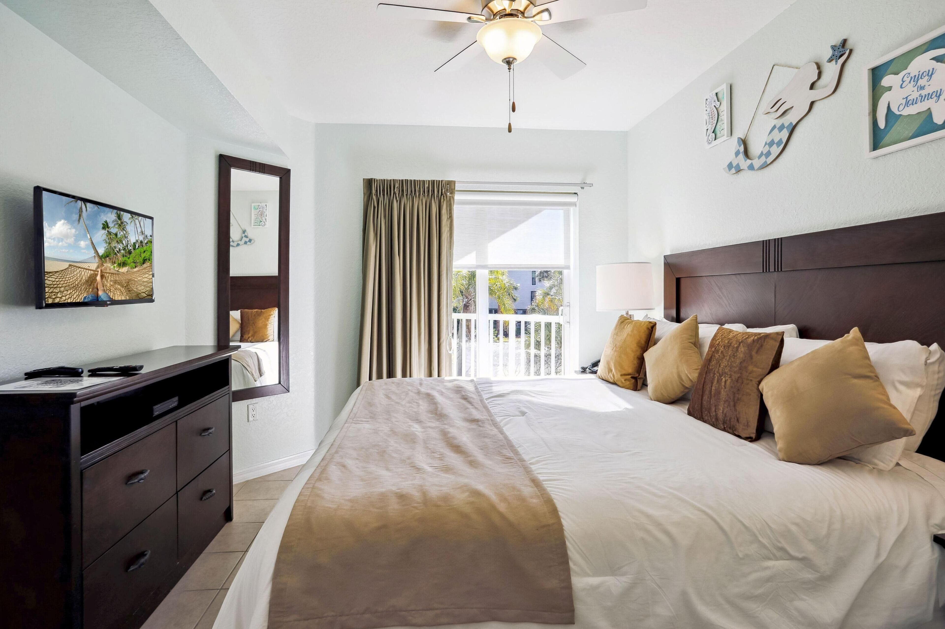 A hotel room with a bed, a TV, a desk, and a balcony with curtains.
