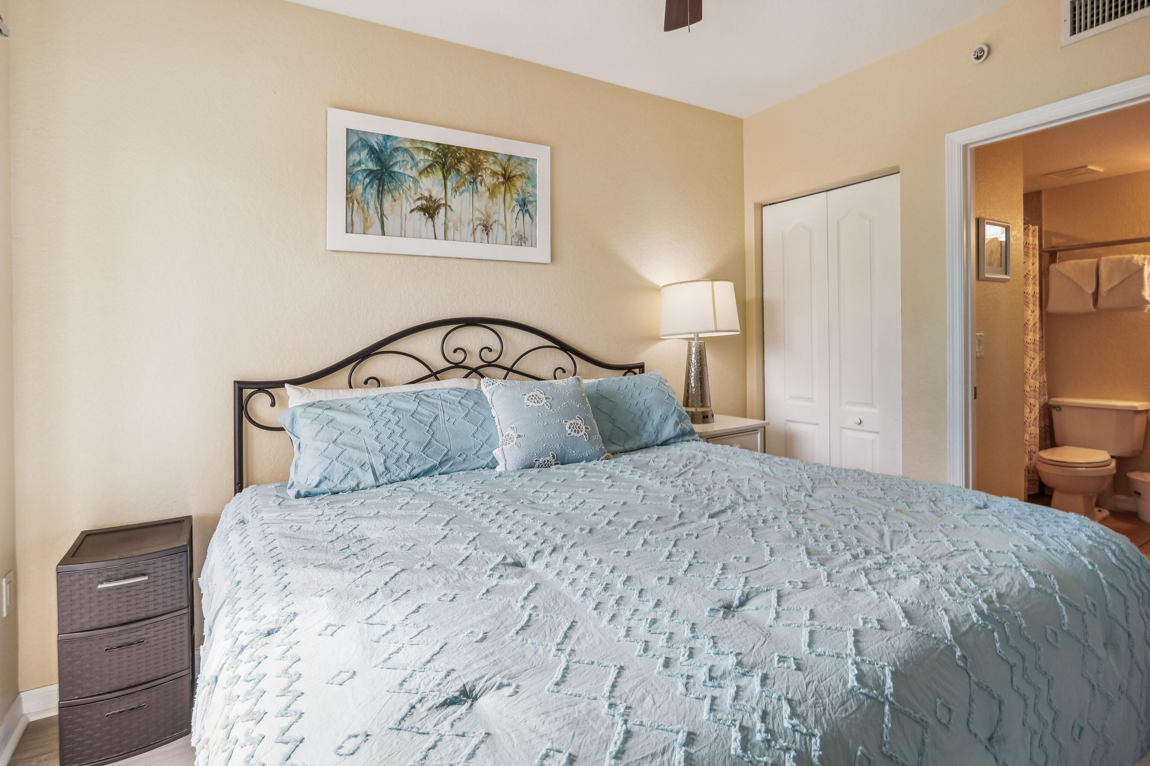 A bedroom with a bed, a nightstand, a drawer unit, a painting, and a bathroom door.