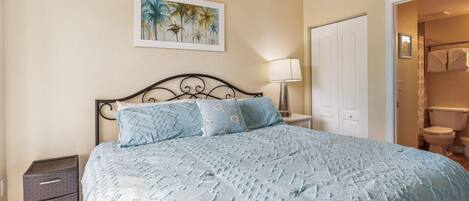 A bedroom with a bed, a nightstand, a drawer unit, a painting, and a bathroom door.