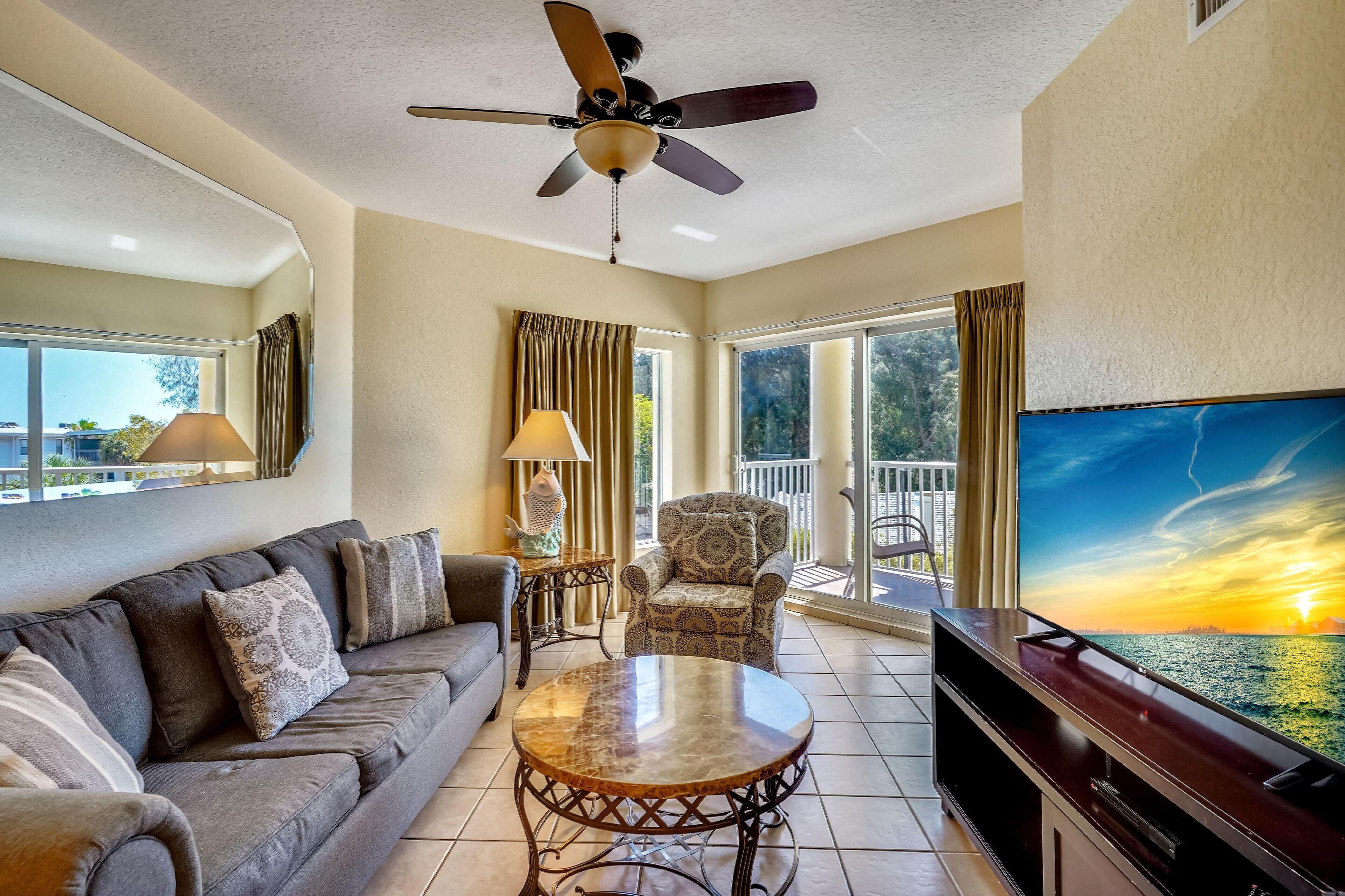 A living room with a sofa, armchair, coffee table, TV, ceiling fan, and large windows with curtains.