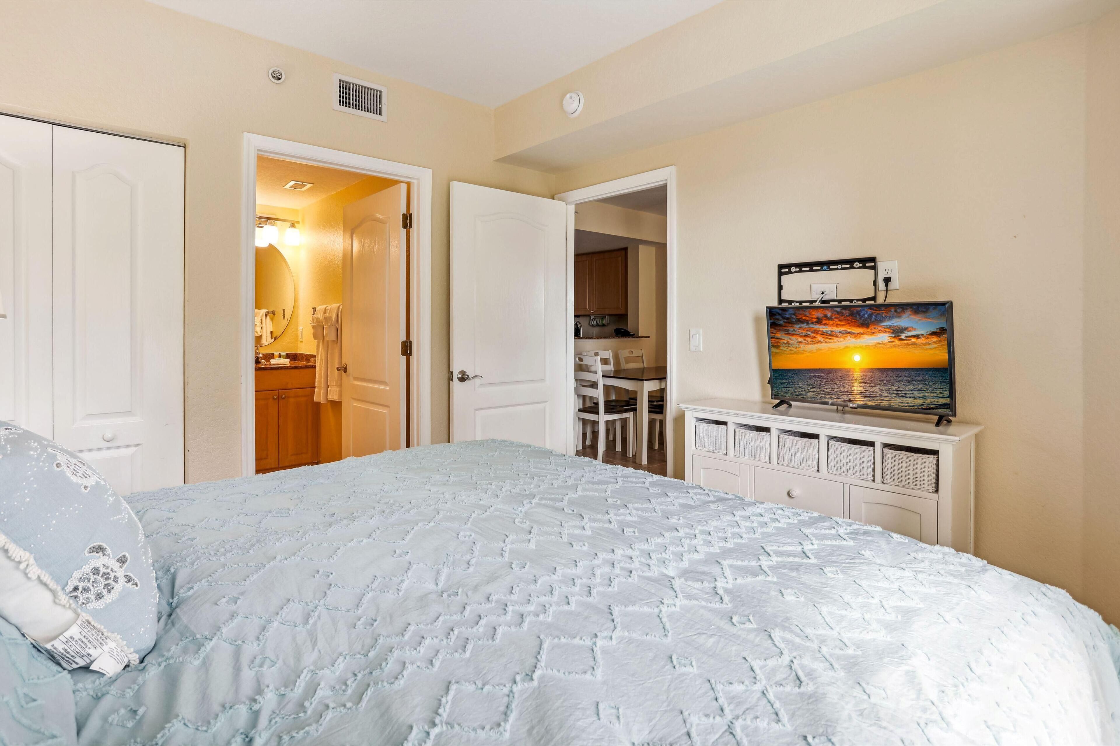 A bedroom with a bed, a TV on a dresser, and a view of the kitchen.