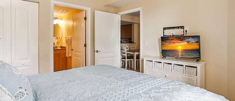 A bedroom with a bed, a TV on a dresser, and a view of the kitchen.