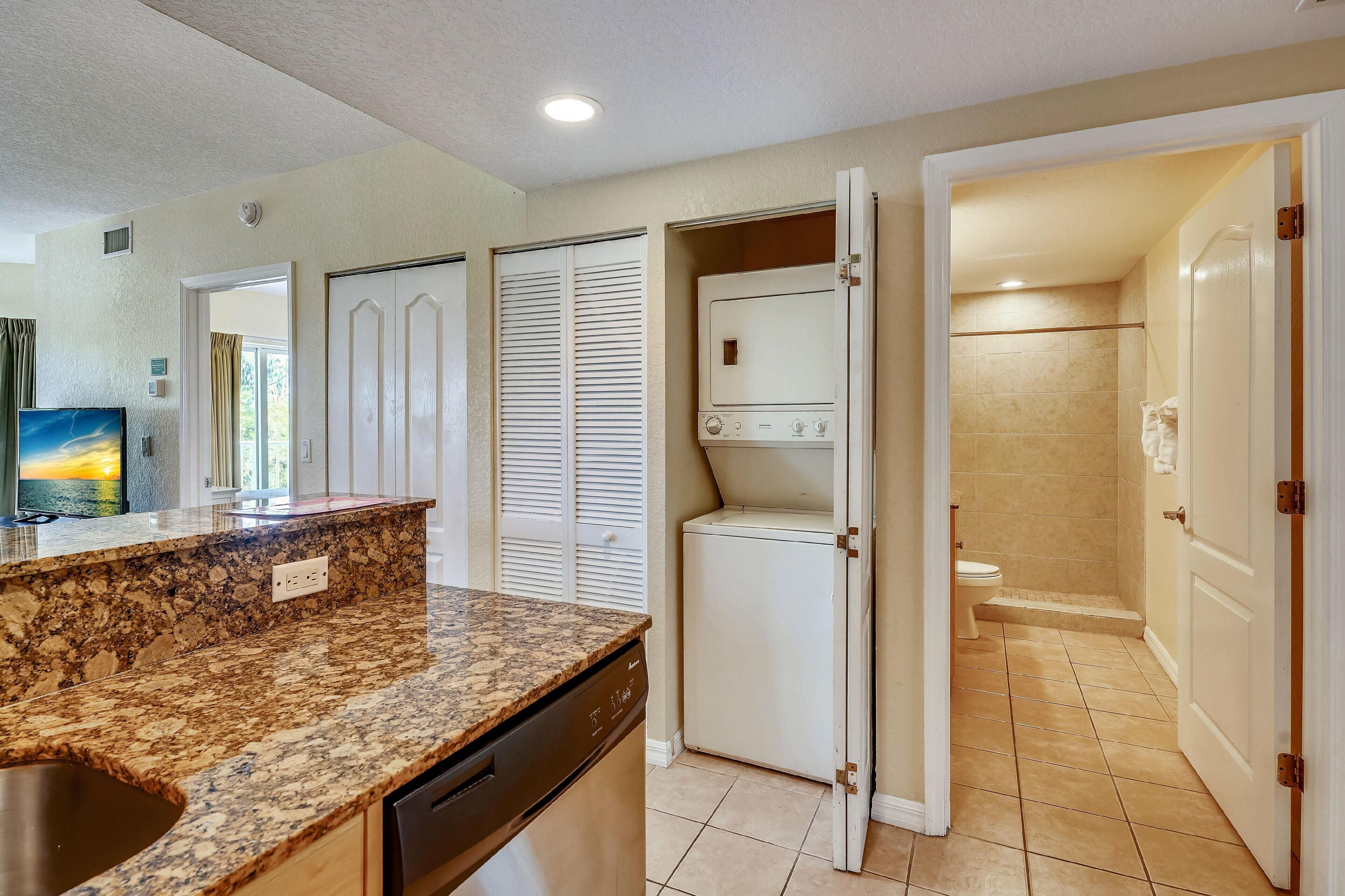 A kitchen with granite countertops, a dishwasher, and a refrigerator. There is a door leading to a bathroom with a shower and toilet.