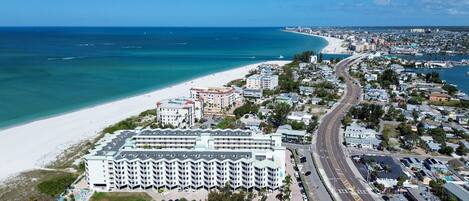 An aerial view of a coastal city with a prominent hotel featuring multiple floors and balconies, a beach, and a busy road.