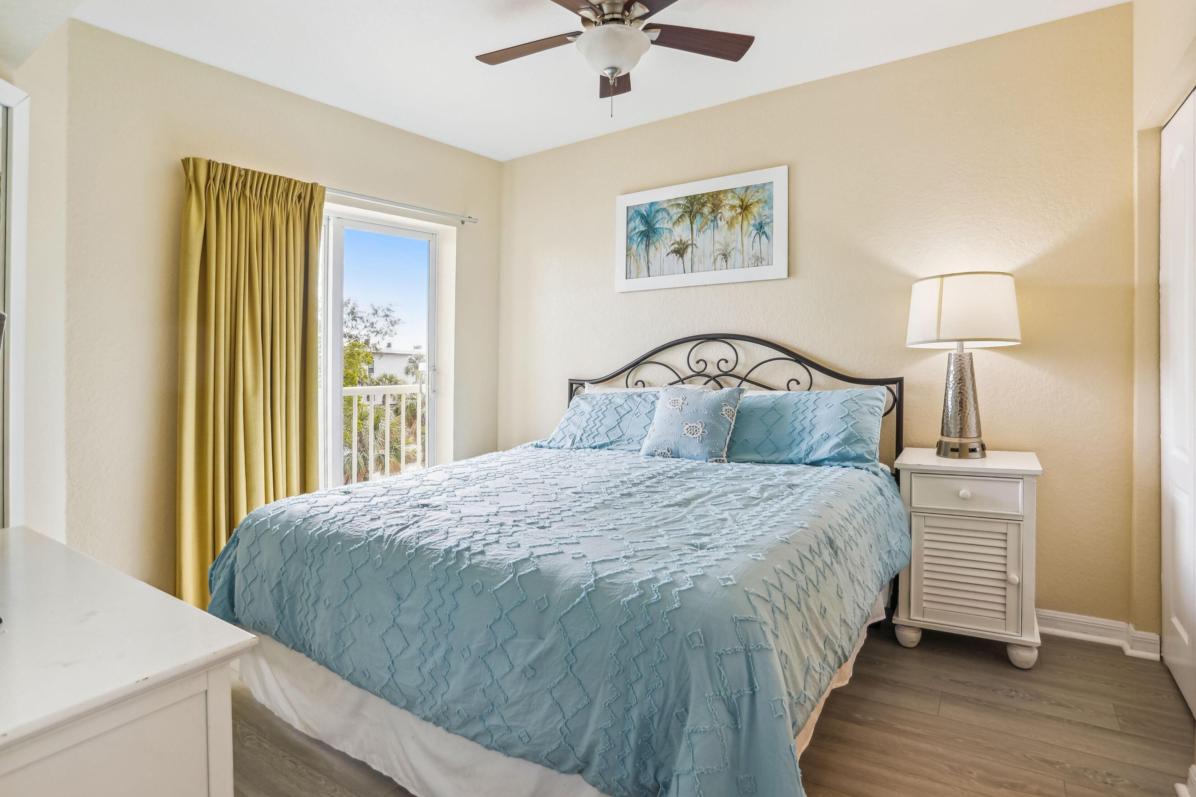 A bedroom with a bed, a nightstand, a ceiling fan, a window with curtains, and a painting on the wall.