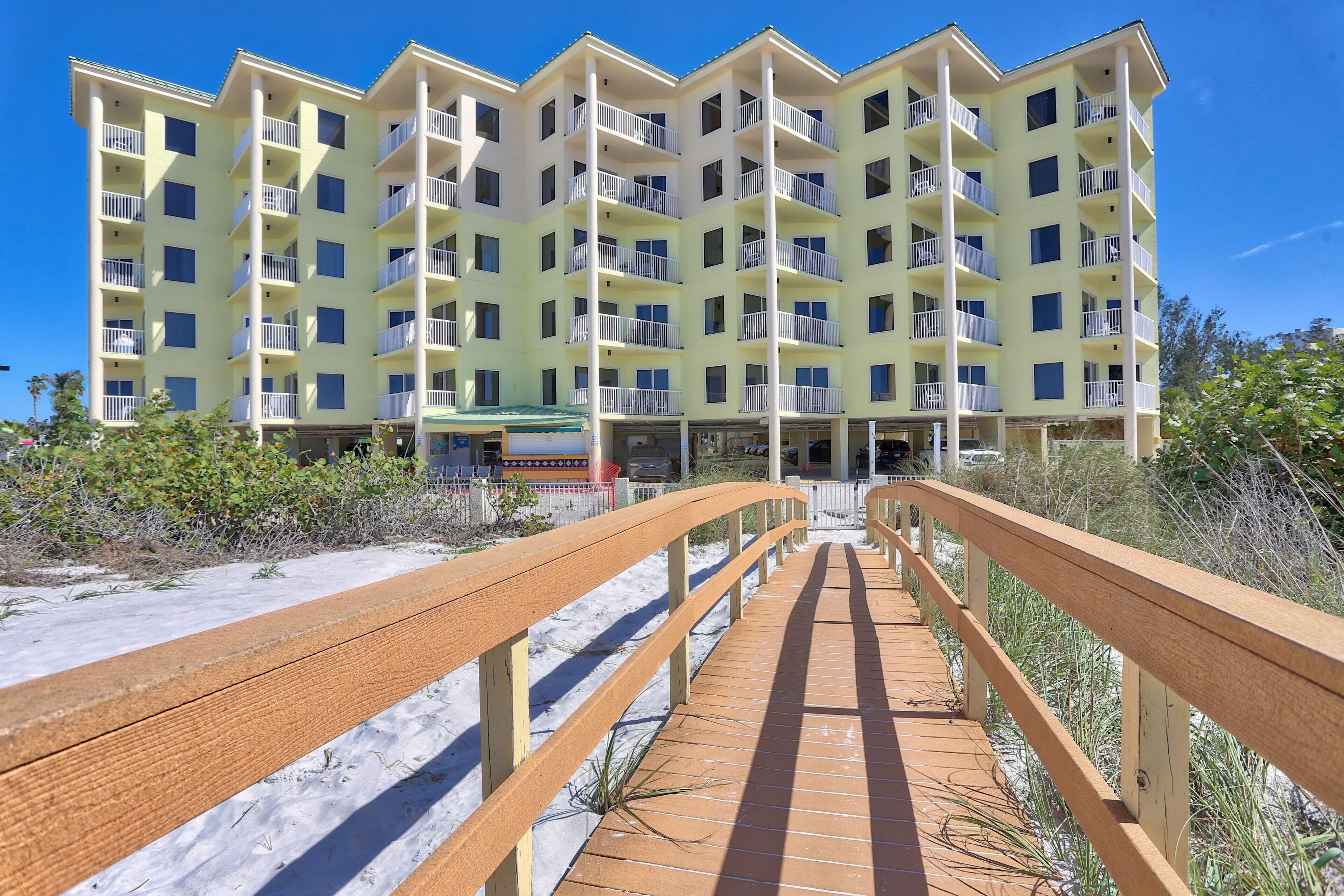 Ein mehrstöckiges Hotel mit Balkonen, ein Sandstrand im Vordergrund und eine Holzpromenade, die zum Gebäude führt.