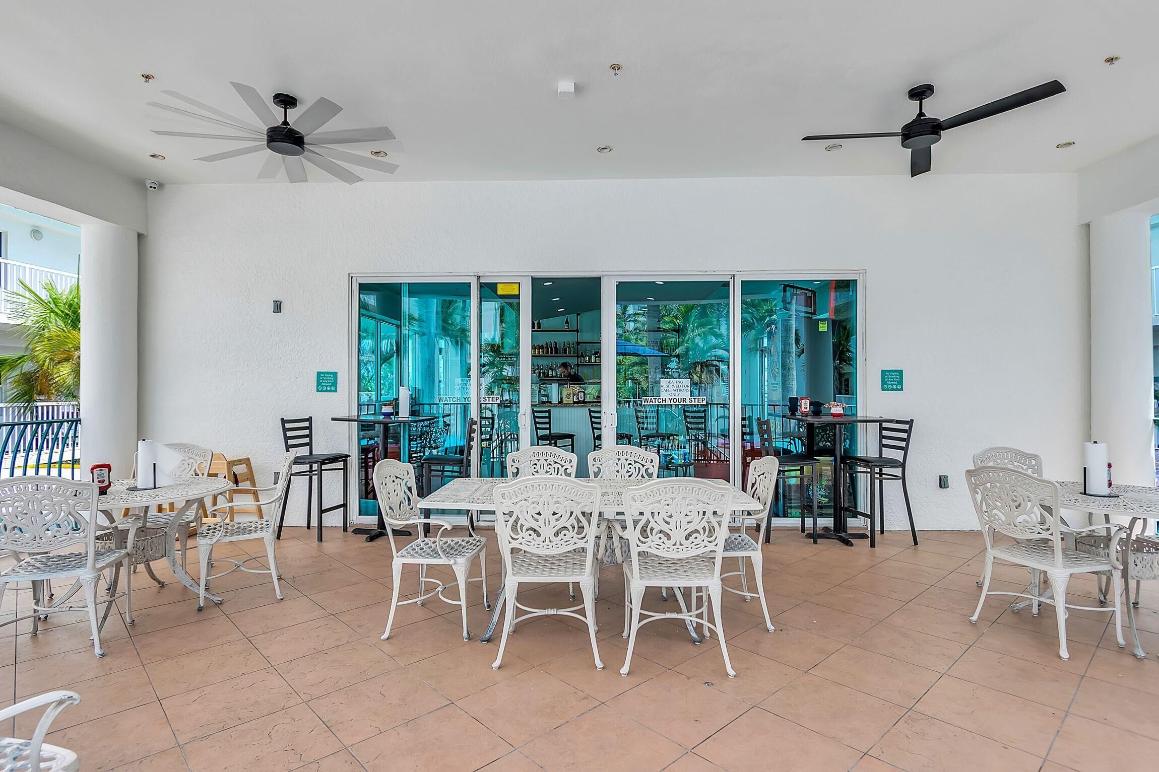 A covered outdoor dining area with white metal furniture, ceiling fans, and a view of the bar area through glass doors.