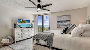 Een slaapkamer met een bed, een plafondventilator, een dressoir, een televisie en een raam met uitzicht op de oceaan.