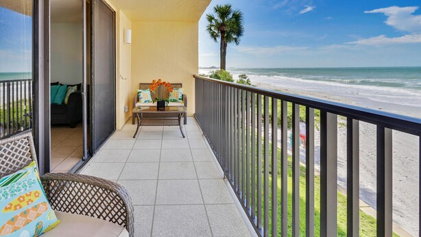 A balcony with a view of the beach, a wicker chair, a table with a vase of flowers, and a sofa with patterned cushions.