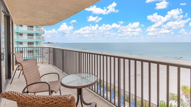 A balcony with wicker chairs and a small table, overlooking a beach and a building with a pool. - Trillium 2A - Premier (Madeira Beach)