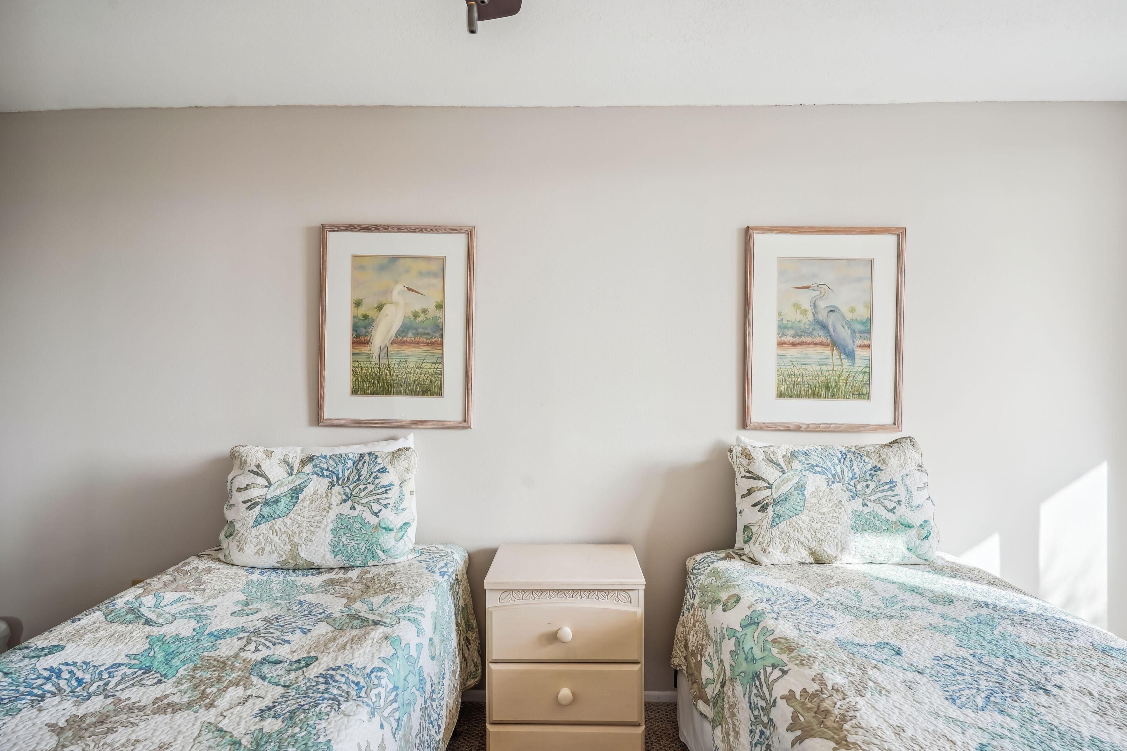 Quarto com duas camas, cada uma com roupa de cama estampada, uma mesinha de cabeceira e duas molduras na parede.