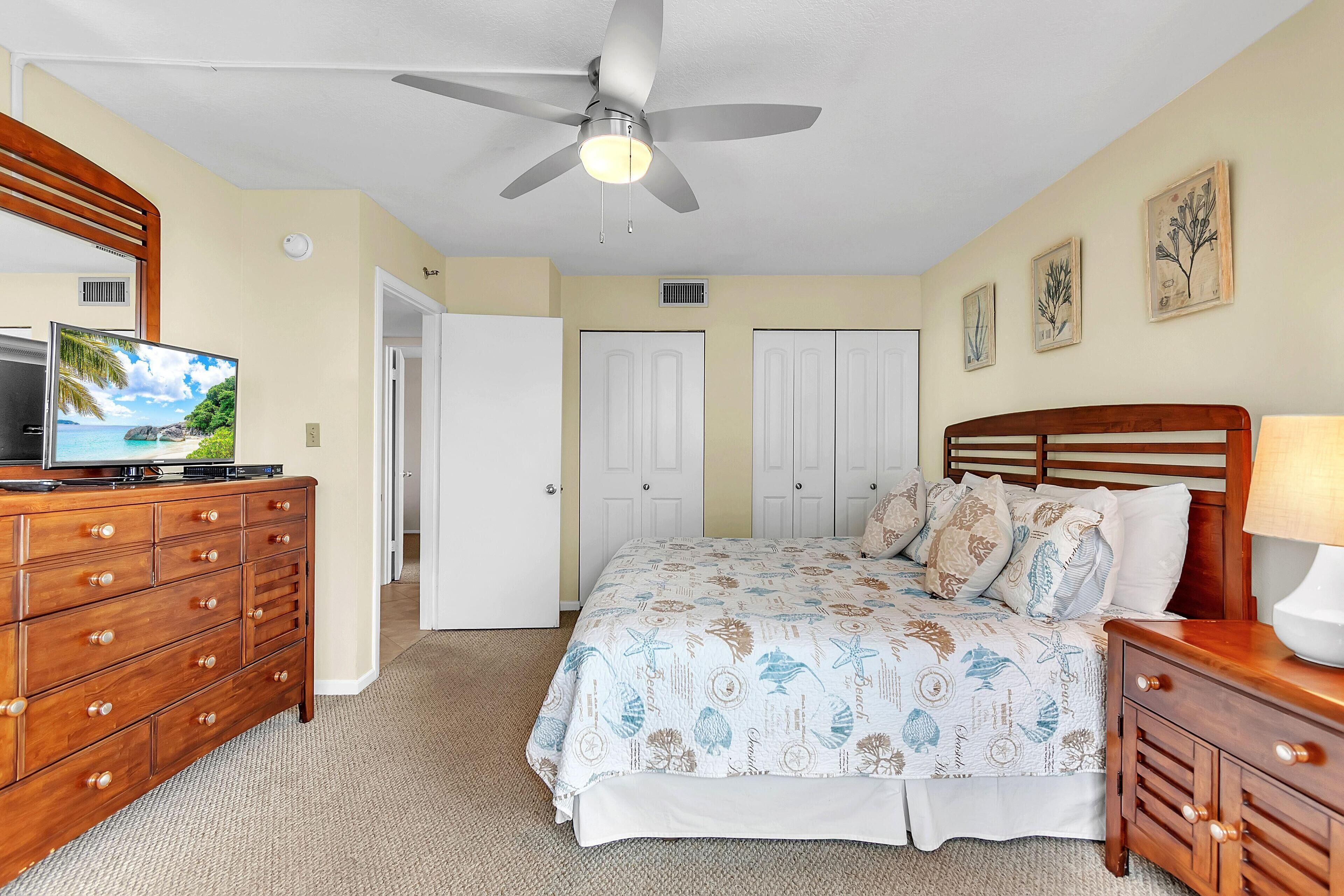 A bedroom with a bed, dresser, TV, ceiling fan, and artwork on the walls.