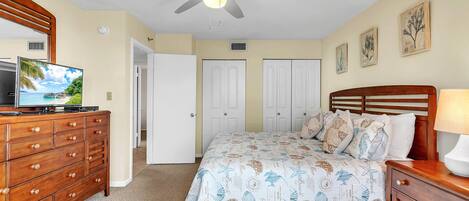 A bedroom with a bed, dresser, TV, ceiling fan, and artwork on the walls.
