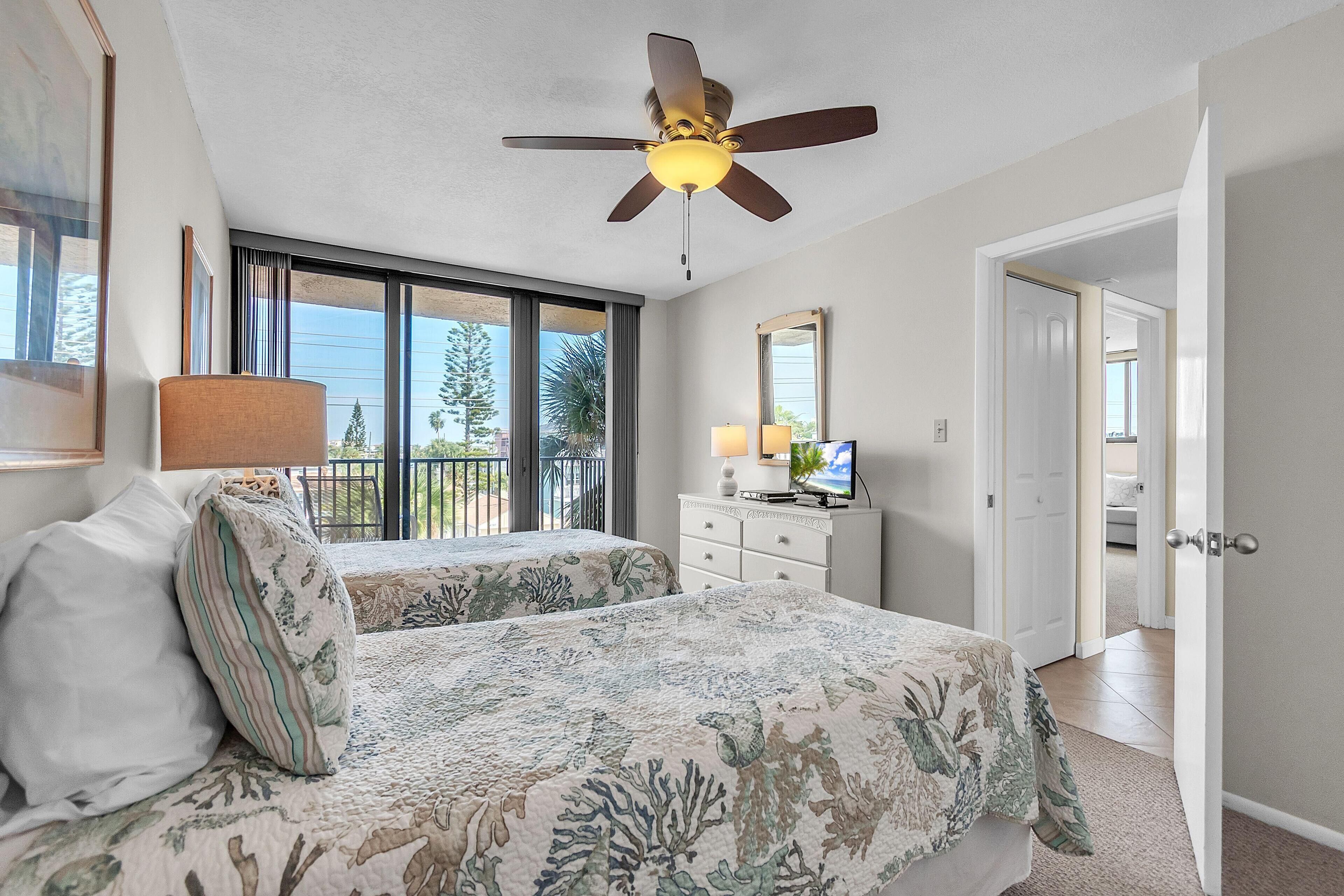 A bedroom with a bed, a ceiling fan, a dresser, a TV, and a balcony with a view of trees.
