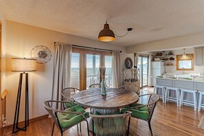 A dining area with a round wooden table surrounded by wicker chairs with green cushions, a pendant light, and a view through large windows.