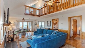 A spacious living area with a large blue sofa, wooden staircase, and a dining area with a table and chairs. - Sun Realty pet-friendly semi-oceanfront home with incredible views! (Kill Devil Hills)