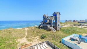 Une maison située en bord de mer, dotée d’une terrasse offrant une vue sur l’océan, d’un chemin de sable menant à la plage et d’un petit espace extérieur aménagé pour s’asseoir.