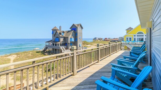 Une terrasse en bois avec des chaises Adirondack bleues, donnant sur la plage, et une maison avec un toit bleu.