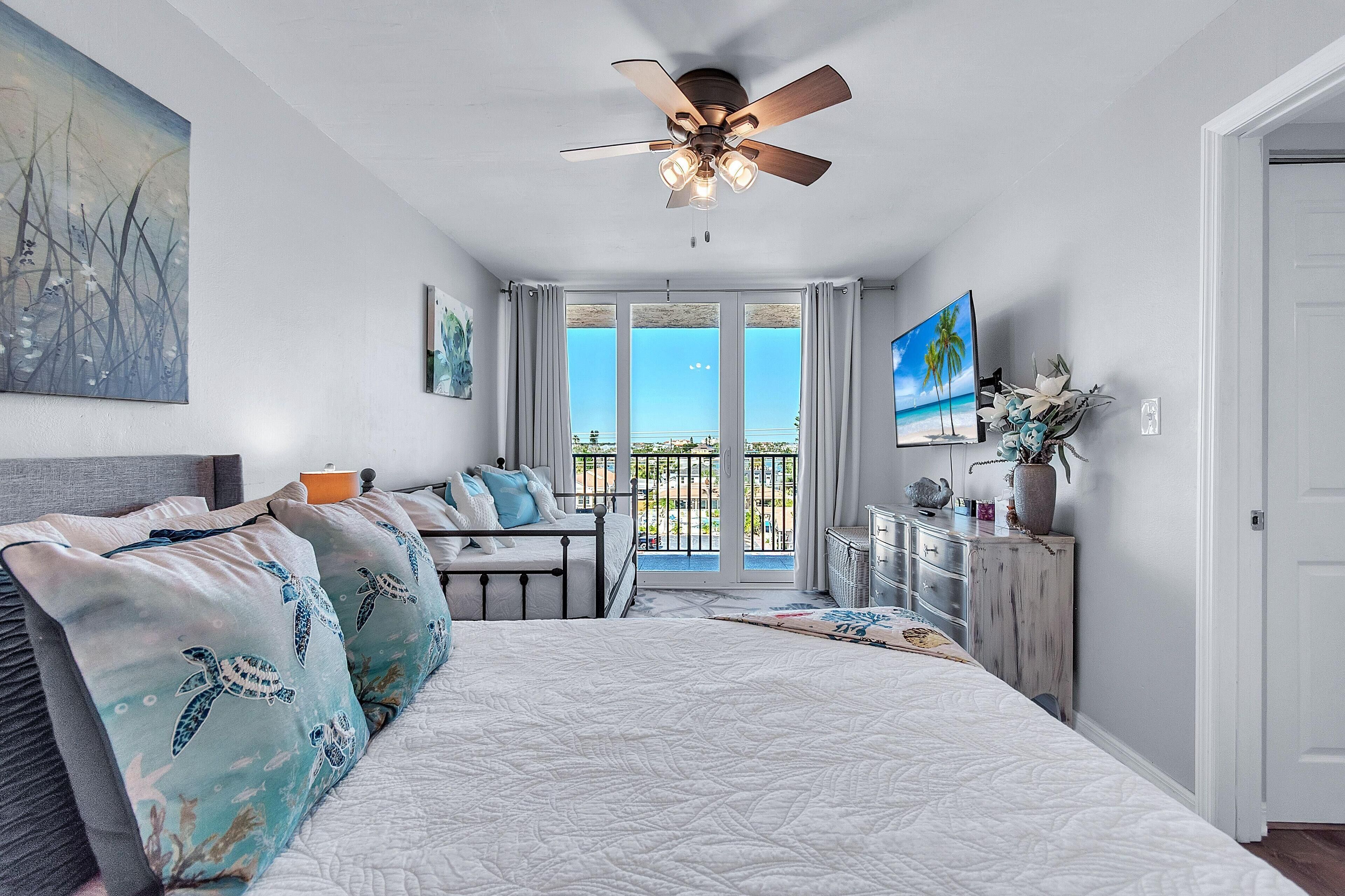 A bedroom with a large bed, a ceiling fan, a view of the outdoors, and a balcony with a railing.