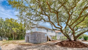 A two-story house with a balcony, surrounded by trees and a sandy yard. - Sun Realty pet-friendly soundside home with private pool! (Kill Devil Hills)