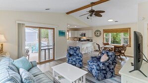 A spacious living room with a sofa, two patterned armchairs, a coffee table, and a television. A kitchen is visible in the background with a dining area and a large window. - Sun Realty pet-friendly soundside home with private pool! (Kill Devil Hills)