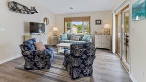 A living room with two patterned armchairs, a sofa, a TV, and a dining area with a table and chairs. - Sun Realty pet-friendly soundside home with private pool! (Kill Devil Hills)