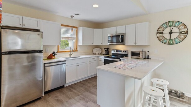 A modern kitchen with stainless steel appliances, white cabinetry, a central island with stools, and a wall clock. - Sun Realty pet-friendly soundside home with private pool! (Kill Devil Hills)