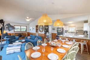 Un vaste espace de vie comprenant une grande table à manger en bois, disposée pour accueillir six personnes, un canapé bleu et, au fond, une cuisine.