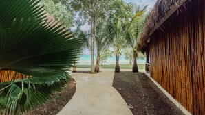 A concrete pathway leading to a wooden structure with a thatched roof, surrounded by tropical vegetation and a view of the beach.