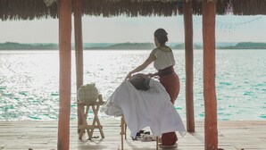 A wooden deck with a thatched roof, a woman sitting on a chair, and a view of the water.